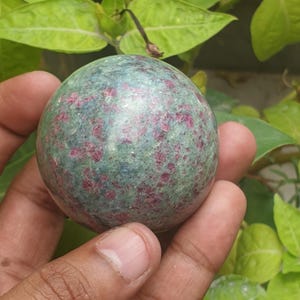 Könnte beinhalten: Eine runde, polierte Edelstein-Kugel. Der Stein ist grün mit roten Einschlüssen und wird in einer Hand gehalten. Im Hintergrund sind grüne Blätter zu sehen.