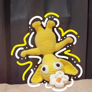 Peut inclure: Un jouet de lapin au crochet jaune avec de grandes oreilles, des yeux noirs et une fleur blanche avec un centre orange. Le lapin est à l'envers. Le fond est un rideau sombre et une surface rose.