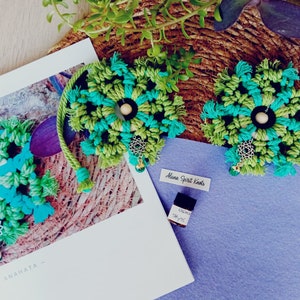 May include: Two green and blue macrame wall hangings with wooden beads and silver charms. The hangings are on a woven straw mat with a small bottle of essential oil labelled "Orange Thyme" and a blue felt square with the text "Aluna Spirit Knots".