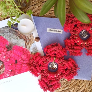 Puede incluir: Dos tapices de pared de forma de flor de color rojo hechos a crochet con un amuleto dorado en el centro. Los amuletos presentan un símbolo que representa el chakra raíz. Los tapices están sobre un fondo morado con el texto "Alana Spirit Knots".