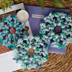 Puede incluir: Posavasos de macramé azul y turquesa hechos a mano con un dije central. Los posavasos están dispuestos sobre una superficie tejida con una pequeña vela y una tarjeta que dice "Alone Spirit Knots" y "VISHUDDHA".