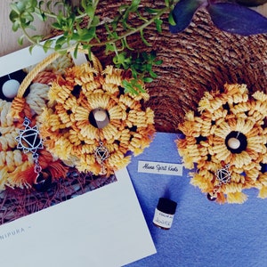 Könnte beinhalten: Zwei gelbe und orangefarbene Makramee-Wandbehänge mit einem silbernen Anhänger in Form eines Dreiecks. Die Wandbehänge befinden sich auf einer blauen Oberfläche mit dem Text "Aluna Spirit Knots".