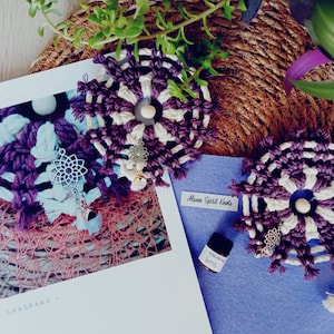 Può includere: Due arazzi in macramè di colore viola e bianco con ciondoli argentati e una piccola bottiglia di olio essenziale etichettata "aroma di eucalipto". Il testo "Aluna Spirit Knots" è scritto su uno sfondo di feltro blu.