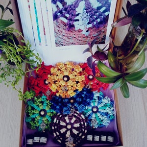 Puede incluir: Un conjunto de siete posavasos de mandala de ganchillo de colores con un amuleto plateado en el centro de cada uno. Los posavasos están dispuestos en círculo sobre un fondo de tela morado. Los posavasos son de color rojo, amarillo, verde, azul y marrón.