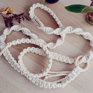 May include: Cream-colored macrame plant hanger with wooden rings and a handle. The handmade design features a woven pattern and a small tag that reads "Home Spirit Life". The plant hanger is displayed on a light wood surface.