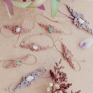 May include: A collection of six macrame bracelets featuring intricate knotted cord patterns in beige and grey tones. Each bracelet is centered with a single polished gemstone, including white, green, and purple stones, accented with small metallic beads.
