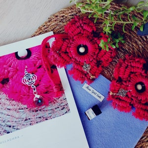 Puede incluir: Dos tapices de pared de flores rojas de ganchillo con un amuleto plateado y una pequeña botella de aceite esencial. El texto "Alana Spirit Knots" es visible en la botella.