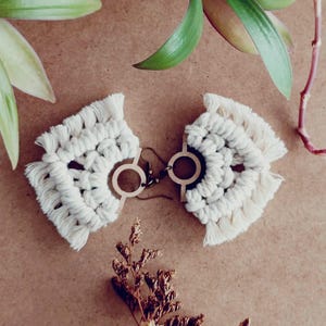 May include: A pair of white macrame earrings with wooden hoops. The earrings have a fringe detail and are hanging from a brown surface.