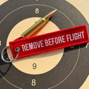 Könnte beinhalten: Ein roter Schlüsselanhänger mit dem Text "REMOVE BEFORE FLIGHT" in weißen Buchstaben. Der Schlüsselanhänger ist an einem goldfarbenen Geschoss befestigt.