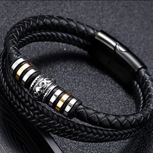May include: A black leather bracelet with a braided design and a silver and gold accent piece. The bracelet has a magnetic clasp.