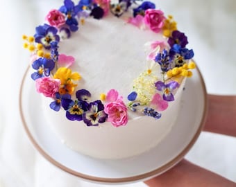 Pode incluir: Um bolo branco com cobertura decorado com uma coroa de flores coloridas. As flores incluem rosas cor-de-rosa, amores-perfeitos roxos, mimosa amarela e lavanda azul. O bolo está sobre um prato branco com borda dourada.
