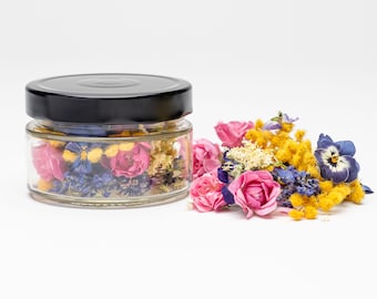May include: A glass jar filled with dried flowers, including pink roses, yellow mimosa, and blue pansies. The jar has a black lid and is sitting on a white surface.