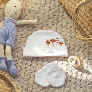 May include: A white baby hat with a mushroom and leaf design and the name "Malo" embroidered on it. The hat is lying on a beige crocheted blanket with a crocheted bunny toy and a wooden teether.