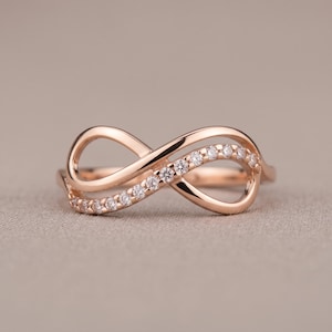 May include: A rose gold infinity ring featuring a polished metal band shaped into a continuous loop. One side of the infinity symbol is encrusted with a row of small, clear, round-cut stones that catch the light, creating a sparkling effect.