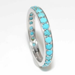 Op de afbeelding: Een zilveren ring met een doorlopende band van kleine, ronde, turkooizen edelstenen. De gepolijste zilveren band van de ring contrasteert met het levendige blauwgroen van de stenen, tegen een witte achtergrond.