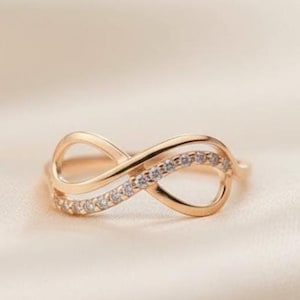 Puede incluir: Un anillo en tono oro rosa con un diseño de símbolo de infinito. Un lado del bucle de infinito es liso y pulido, mientras que el otro lado está incrustado con una hilera de pequeñas piedras claras y brillantes que captan la luz.