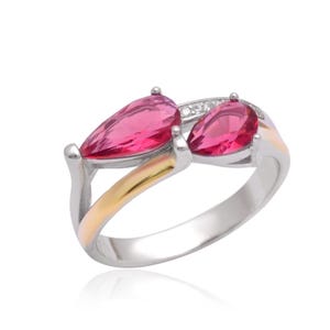 May include: A silver ring with two pear-shaped, bright pink gemstones. The ring has gold accents and small, clear stones set between the pink gems. The design is modern and elegant, suitable for a special occasion.