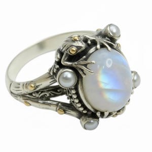 Op de afbeelding: Zilveren ring met een ovale, iriserende maansteen. De ring is versierd met een kikker en kleine parels. De band heeft een bamboe-achtig ontwerp met gouden accenten. Een uniek sieraad.