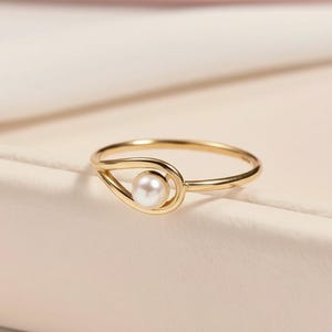 May include: A delicate gold ring with a small, round, white pearl. The ring's design includes a teardrop-shaped setting that holds the pearl, with a slender band. The ring is displayed on a neutral-toned surface.