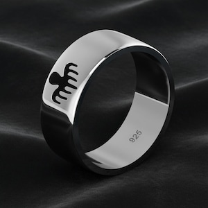 Peut inclure: Bague argentée avec un emblème noir sur le dessus. La bague est en métal poli et porte le numéro "925" gravé à l'intérieur. L'anneau est présenté sur un fond sombre.