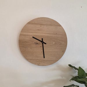 May include: A round wooden clock with black hands on a white wall. The clock is made of light wood with a natural grain pattern.
