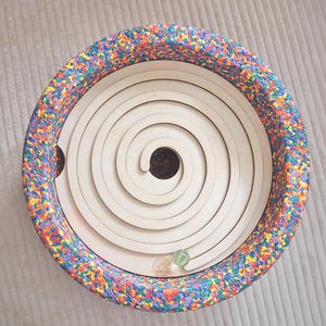 Könnte beinhalten: Ein Holz-Spiral-Labyrinth-Spielzeug mit einem bunten, gesprenkelten Rand. Das Labyrinth weist eine Reihe konzentrischer kreisförmiger Bahnen auf, die zu einem zentralen Loch führen. Eine klare Murmel befindet sich auf der Bahn. Das Wort "HERRLICH" ist auf der Seite aufgedruckt.