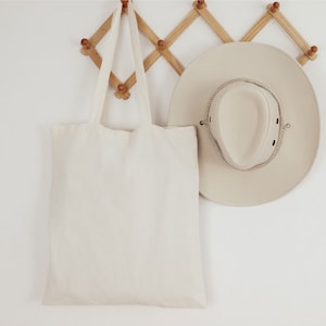 May include: A plain white canvas tote bag hanging on a wooden rack with a beige cowboy hat next to it.