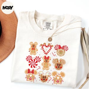 A white t-shirt with a graphic design featuring various Disney characters, including Minnie Mouse, Mickey Mouse, and other popular characters, along with gingerbread cookies and candy.