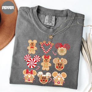 A gray t-shirt with a graphic design featuring various cartoon characters, including Mickey Mouse, Minnie Mouse, and other popular Disney characters, along with gingerbread houses and other festive elements.