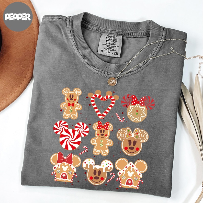 A gray t-shirt with a graphic design featuring various cartoon characters, including Mickey Mouse, Minnie Mouse, and other popular Disney characters, along with gingerbread houses and other festive elements.