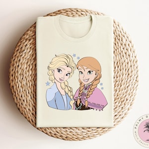 May include: A cream-colored t-shirt with a cartoon image of two girls with long blonde hair. The girls are wearing blue and purple dresses and have blue eyes. The text "Palmy Dreams Co." is printed on the bottom right corner of the shirt.