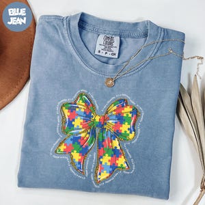May include: A blue Comfort Colors t-shirt featuring a colorful puzzle piece bow design. The bow is outlined in gold and surrounded by the text "It's not a disability, it's a different ability." A gold necklace rests on the shirt.