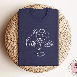 May include: A navy blue t-shirt with a white outline of Mickey Mouse in a playful pose. The design features Mickey Mouse with his arms outstretched and a playful expression on his face.