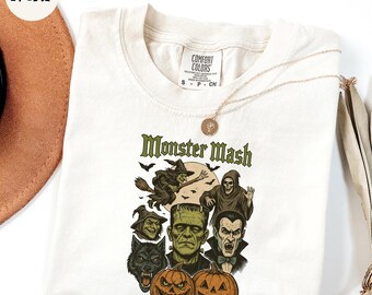 May include: Ivory-colored t-shirt featuring a Halloween-themed graphic with the text "Monster Mash." The design includes illustrations of a witch on a broom, a grim reaper, Frankenstein's monster, a werewolf, and pumpkins. A gold necklace is draped over the shirt.
