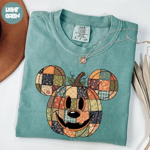 May include: A light green Comfort Colors t-shirt featuring a patchwork pumpkin design in the shape of a mouse. The pumpkin has a smiling face and is made of various fall-themed fabrics. A silver necklace is draped across the shirt.