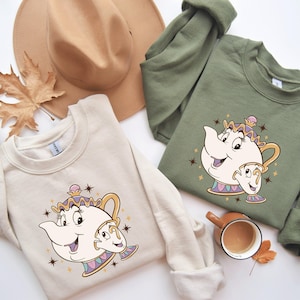 May include: Two cream colored sweatshirts with a cartoon image of a teapot and a teacup. The teapot has a purple lid and a pink face. The teacup has a yellow face. The sweatshirts are on a white background with a brown hat and a leaf.