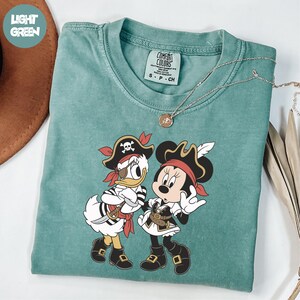 A turquoise t-shirt with a pirate-themed graphic of Minnie Mouse and Donald Duck printed on it, along with a necklace and a brown hat.