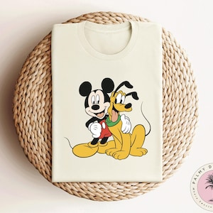 May include: A white t-shirt with a cartoon image of Mickey Mouse and Pluto. Mickey Mouse is wearing red shorts and Pluto is wearing a red collar. The image is centered on the shirt.