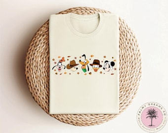 May include: A white t-shirt with a line of fall-themed Disney characters. The characters include Goofy, Pluto, Donald Duck, and Mickey Mouse. The characters are wearing fall-themed outfits and have autumn leaves around them.