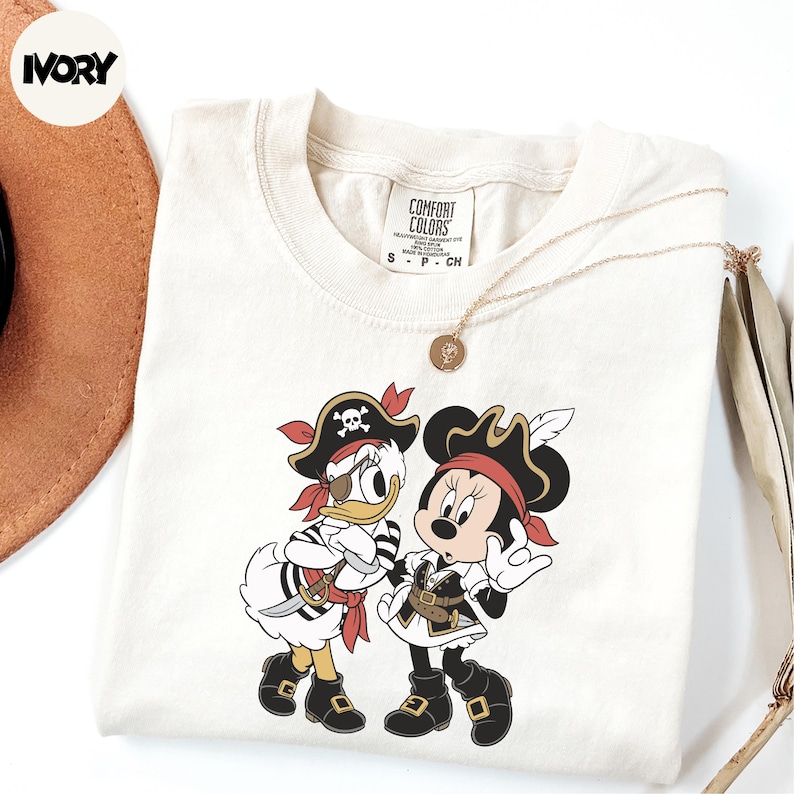 A white t-shirt with a cartoon image of Minnie Mouse and Donald Duck dressed as pirates, along with a necklace and a brown hat.