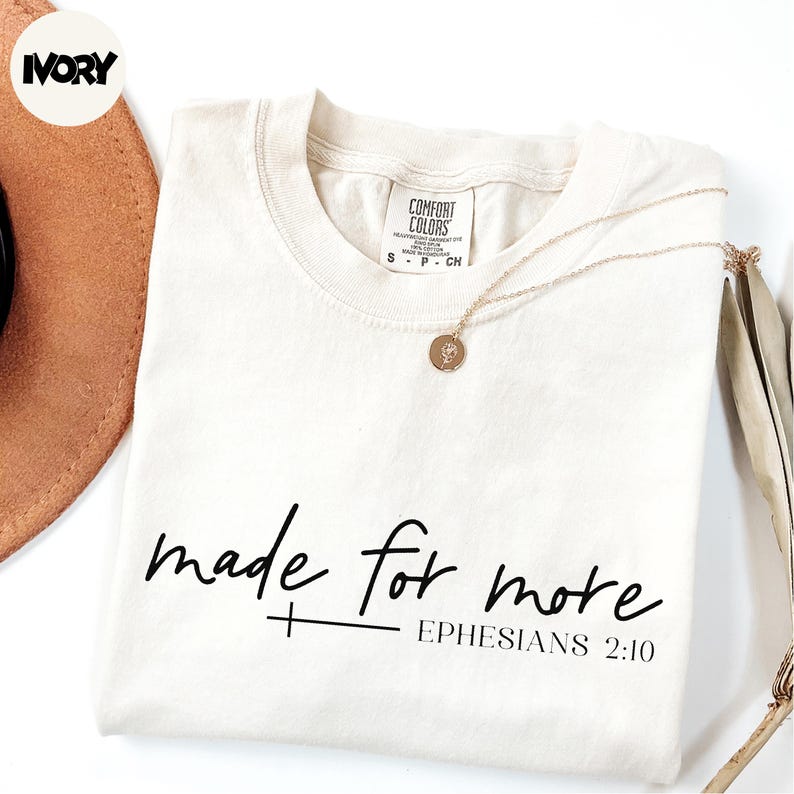 A white t-shirt with the text "made for more" printed on it, along with a small cross symbol and the Bible verse "Ephesians 2:10". The t-shirt is laid out on a white background, and there is a brown hat and a necklace visible in the image.