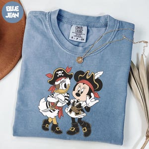 A blue t-shirt with a cartoon image of Minnie Mouse and Donald Duck dressed as pirates, along with a necklace and a brown hat.