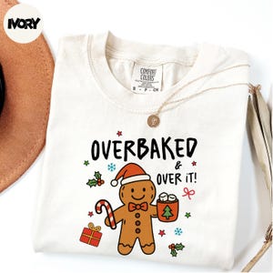 May include: Cream-colored sweatshirt with the text "OVERBAKED & OVER IT!" and a gingerbread man graphic wearing a Santa hat. The graphic includes a candy cane, gift, and a cup of marshmallows. A gold necklace is draped across the shirt.