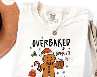 May include: Cream-colored sweatshirt with the text "OVERBAKED & OVER IT!" and a gingerbread man graphic wearing a Santa hat. The graphic includes a candy cane, gift, and a cup of marshmallows. A gold necklace is draped across the shirt.