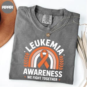 May include: A gray t-shirt with the words "LEUKEMIA AWARENESS" above a rainbow and orange ribbon graphic. The text "WE FIGHT TOGETHER" is below the graphic. A silver necklace with a pendant is on the shirt.