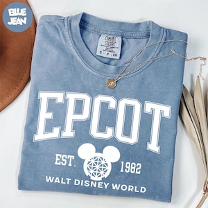 May include: A blue Comfort Colors t-shirt with the word "EPCOT" in white block letters. Below the word "EPCOT" is "EST. 1982" and the words "WALT DISNEY WORLD". A gold necklace with a pendant is on the shirt.