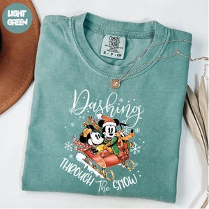 May include: A pale green t-shirt featuring the words "Dashing Through The Snow" and a festive graphic of Mickey, Minnie, and Pluto on a sleigh. The top has a crew neck and short sleeves.