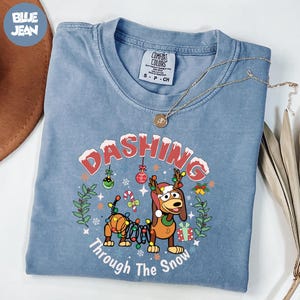May include: A blue Comfort Colors t-shirt featuring the text "Dashing Through The Snow" and a cartoon dog wearing a reindeer antler hat. The shirt has a crew neck and short sleeves. A gold necklace is draped across the shirt.