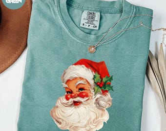 May include: A light green t-shirt featuring a vintage Santa Claus graphic. The Santa illustration has a red hat, white beard, and holly accents. The shirt has a crew neck and short sleeves. A silver necklace with a pendant is draped across the shirt.