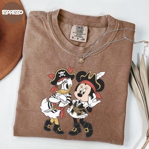 A brown t-shirt with a cartoon image of Minnie Mouse and Donald Duck dressed as pirates, along with a necklace and a hat.